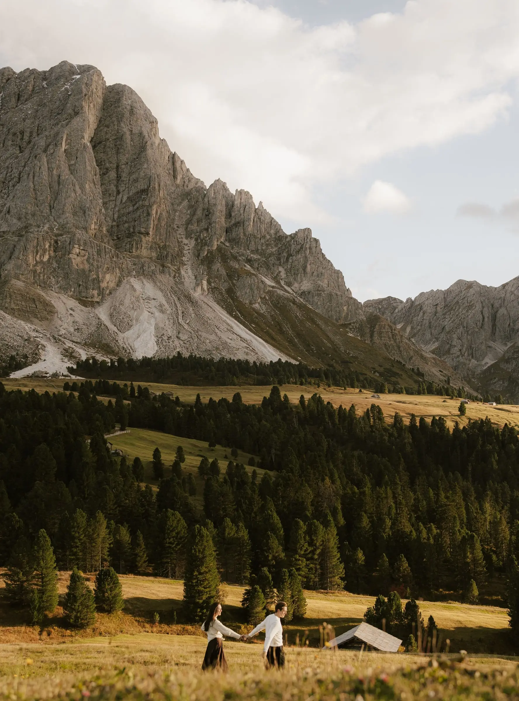 Couple holding hands in a golden meadow below the rock walls of Passo delle Erbe