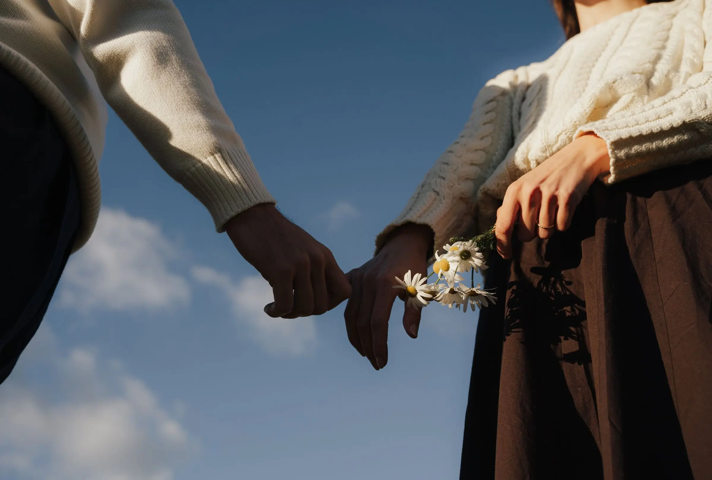 Close-up of the couple holding hands, one holding daisies, against a blue sky