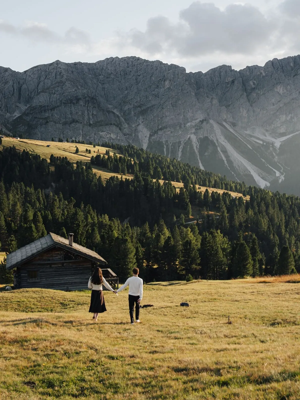 Couple walking across a golden meadow towards wooden huts in the Dolomites