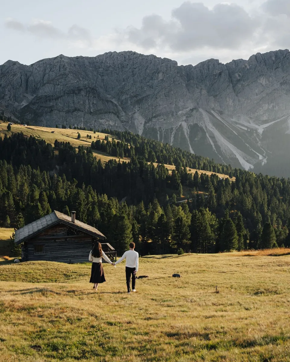 Couple walking across a golden meadow towards wooden huts in the Dolomites