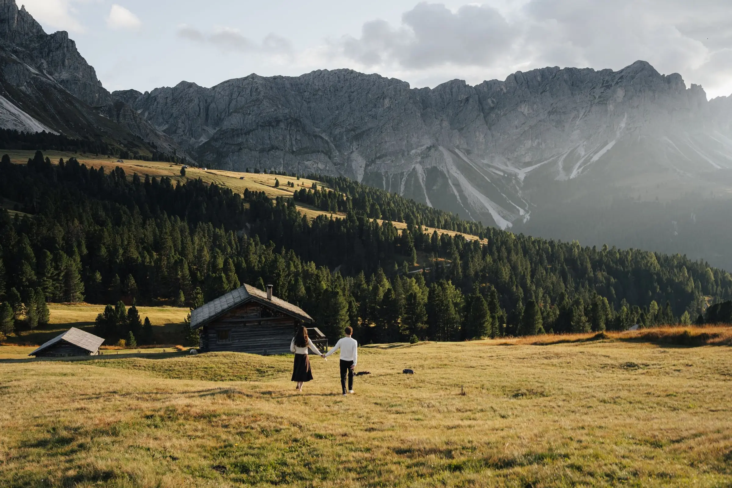 Couple walking across a golden meadow towards wooden huts in the Dolomites