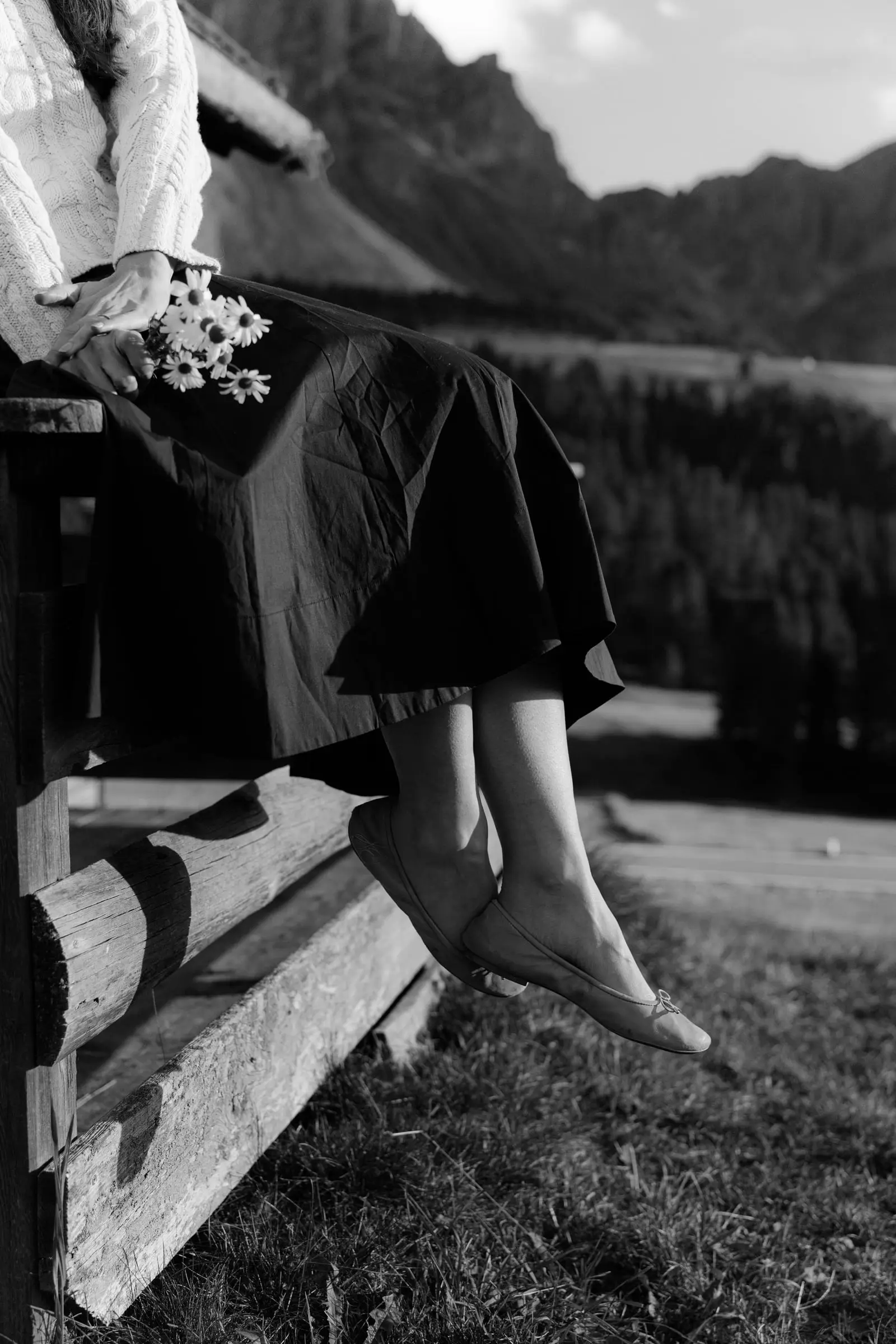 Black and white detail of her feet dangling from a wooden fence, daisies in hand