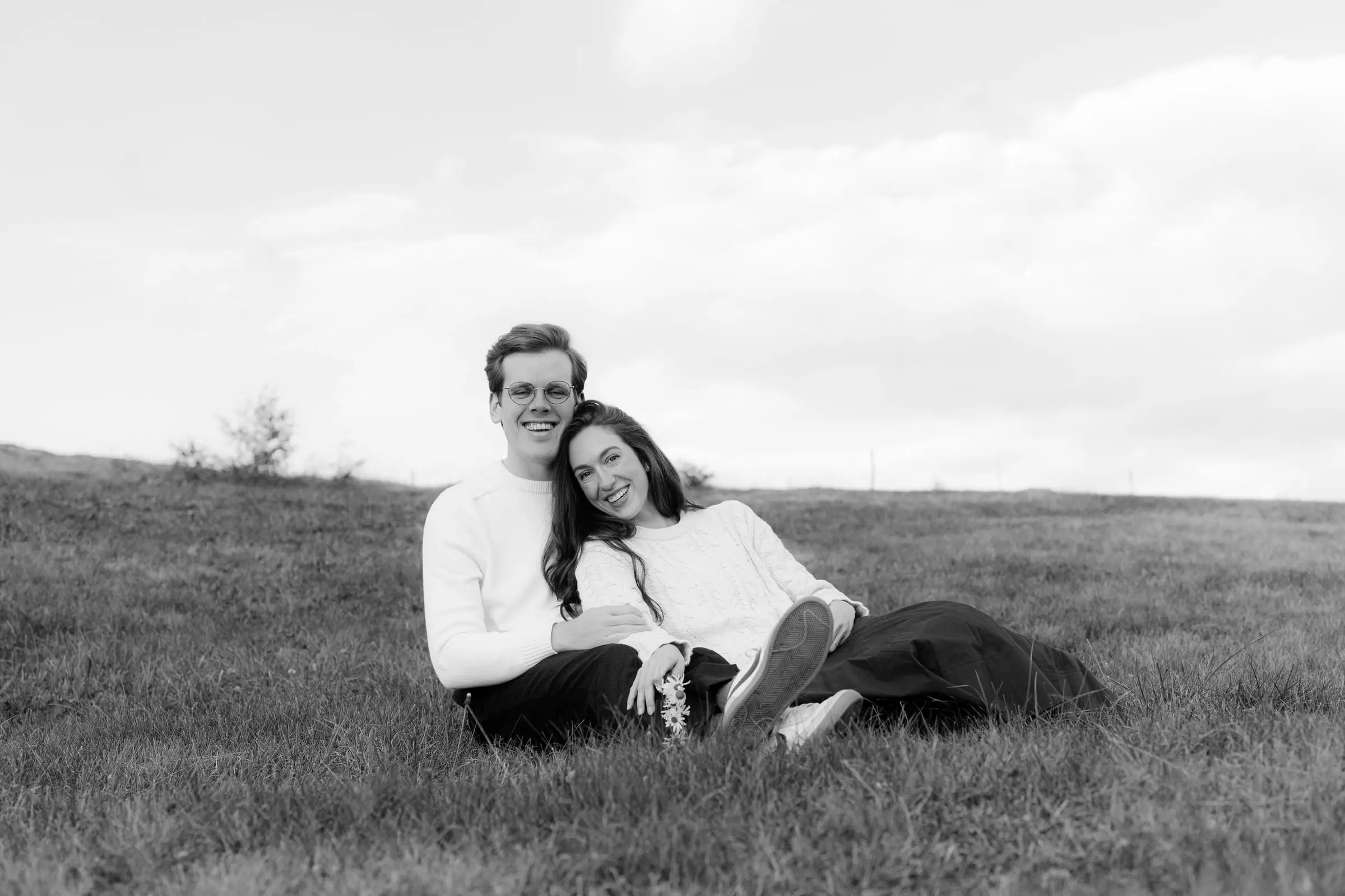Black and white photo of the couple sitting in the meadow, leaning together