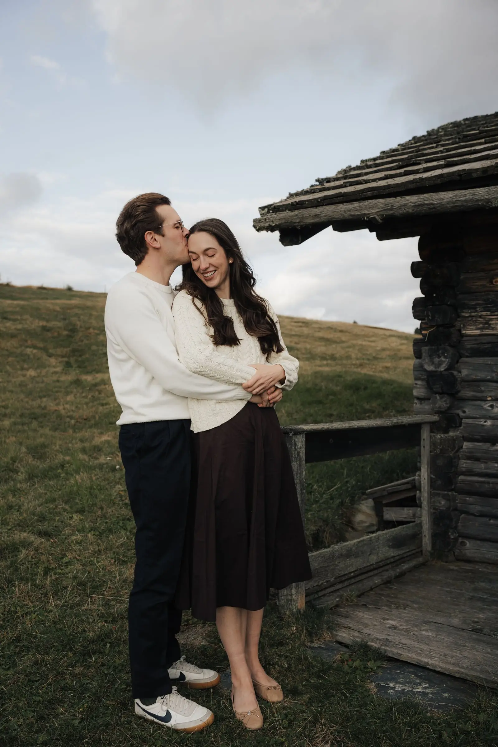 He kisses her forehead next to a wooden hut