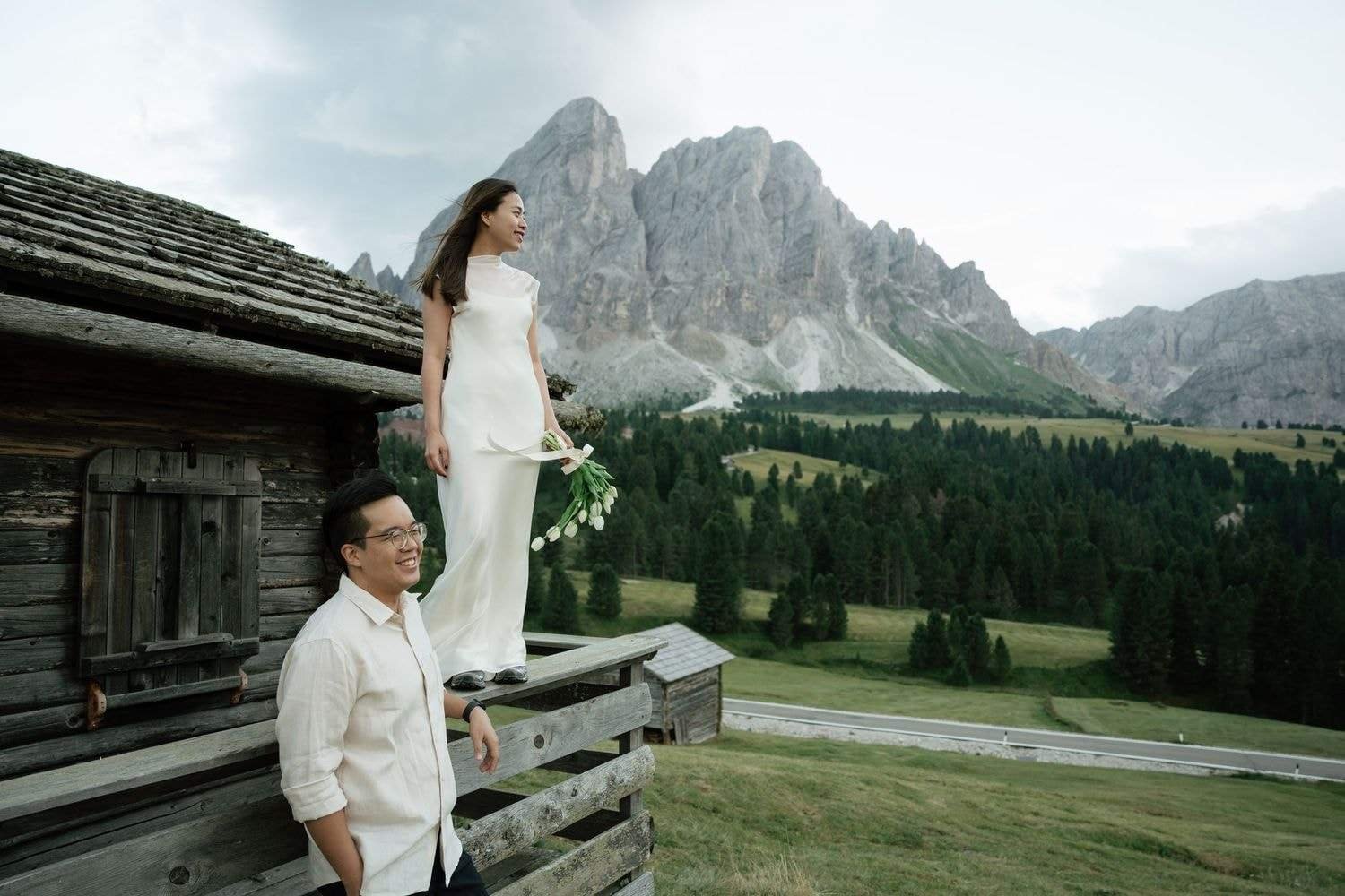 She stands on the fence of a wooden hut in a white dress, he laughs below, Sass de Putia behind