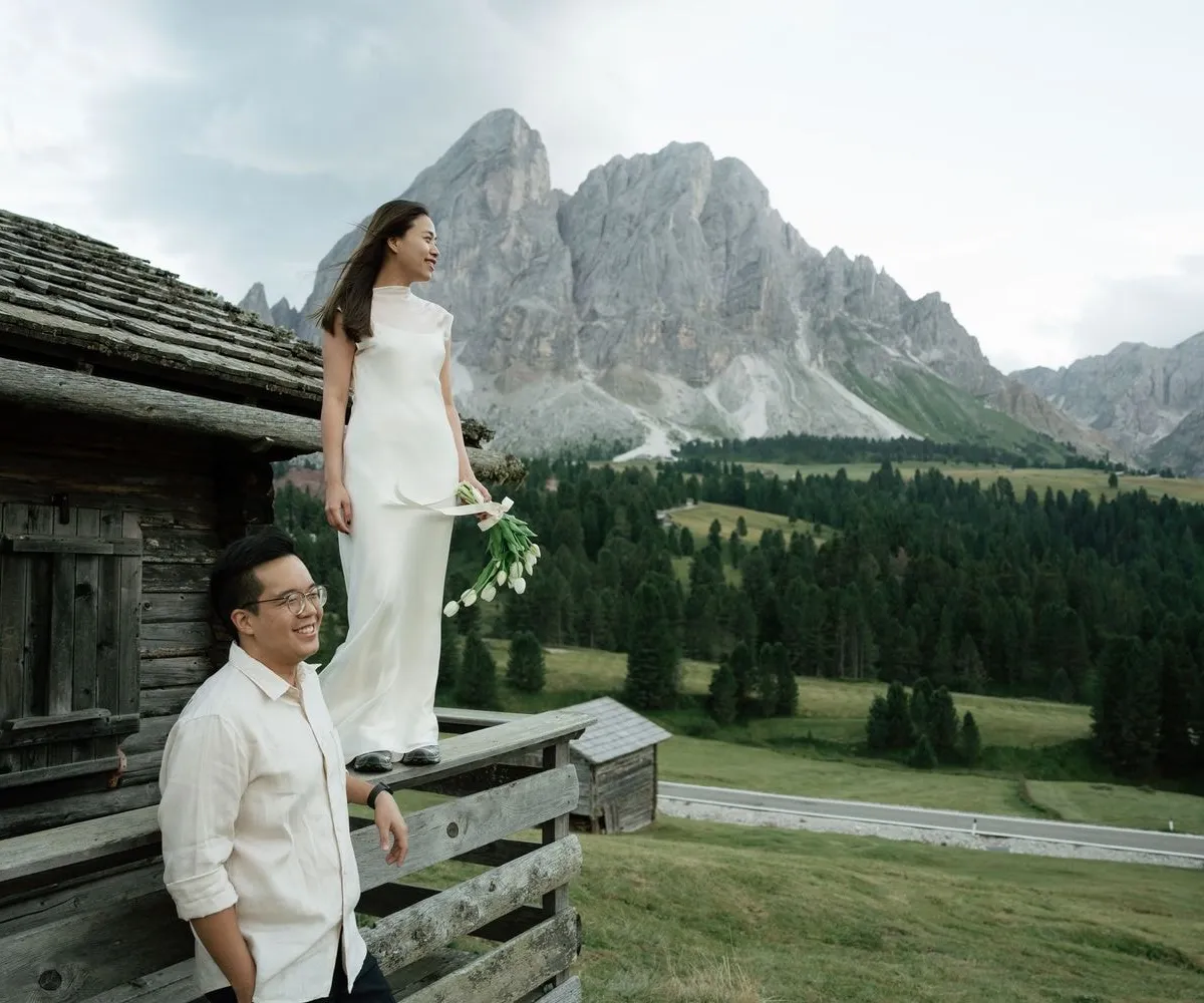 She stands on the fence of a wooden hut in a white dress, he laughs below, Sass de Putia behind