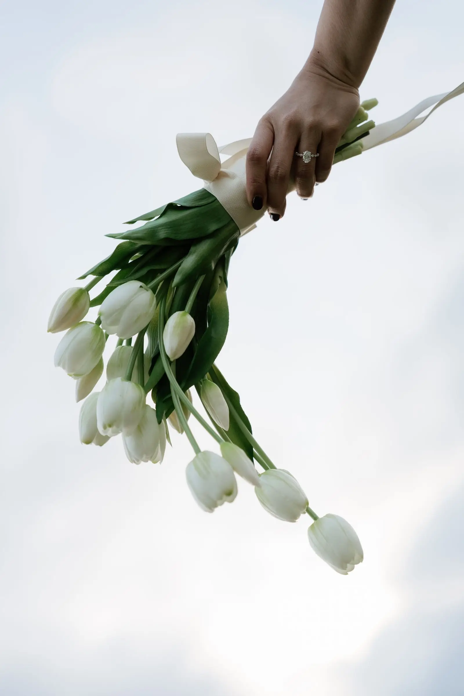 Hand with an engagement ring holding a bouquet of white tulips against the sky