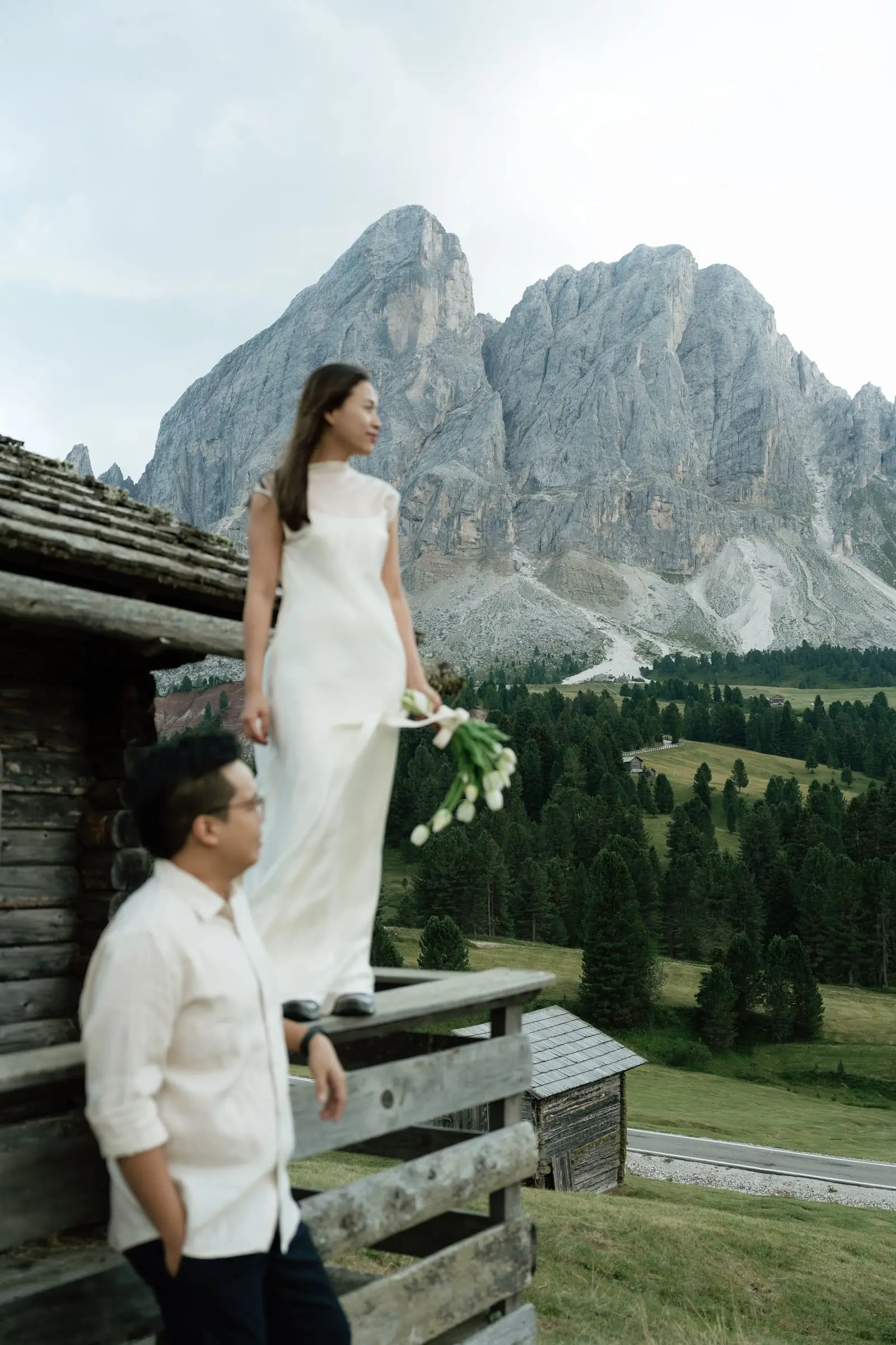 She stands on a hut railing holding white tulips, he leans on the wall