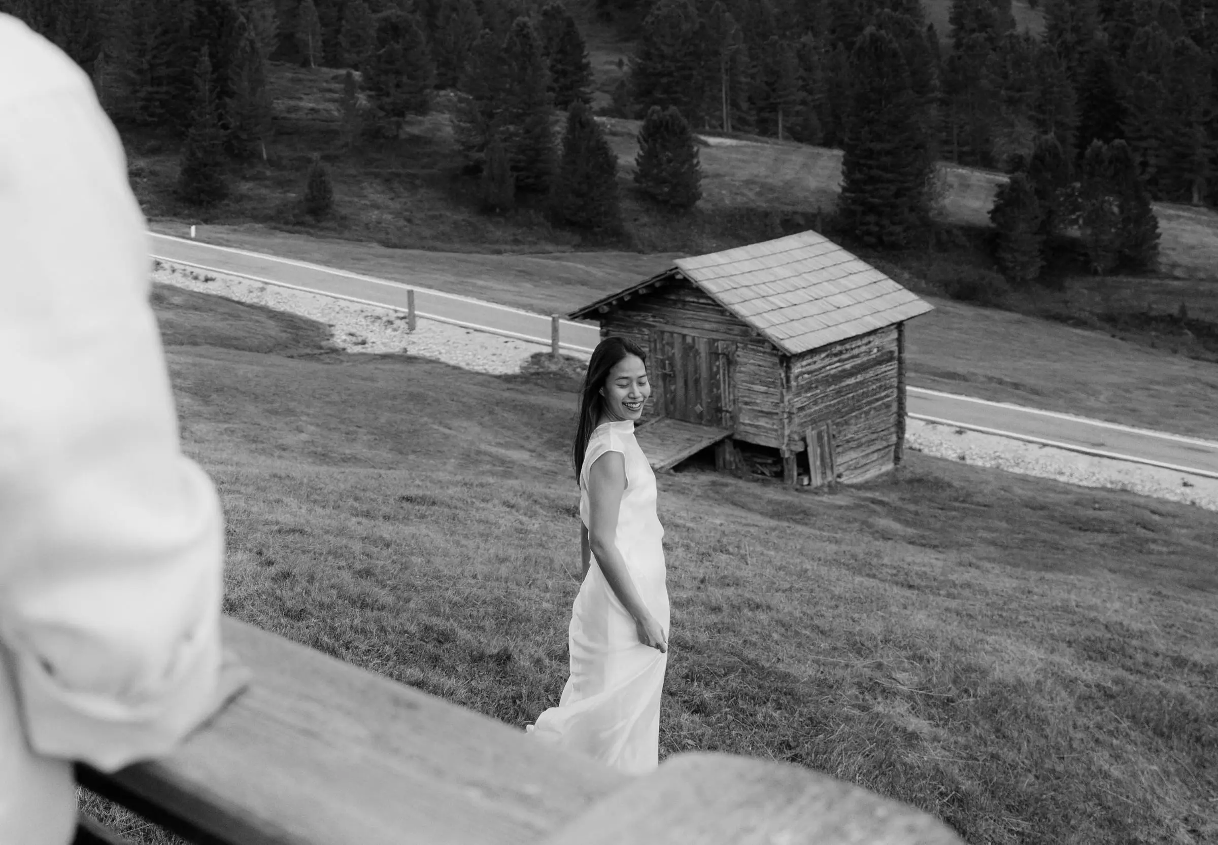 Black and white photo of her in a white dress in the meadow near a hut