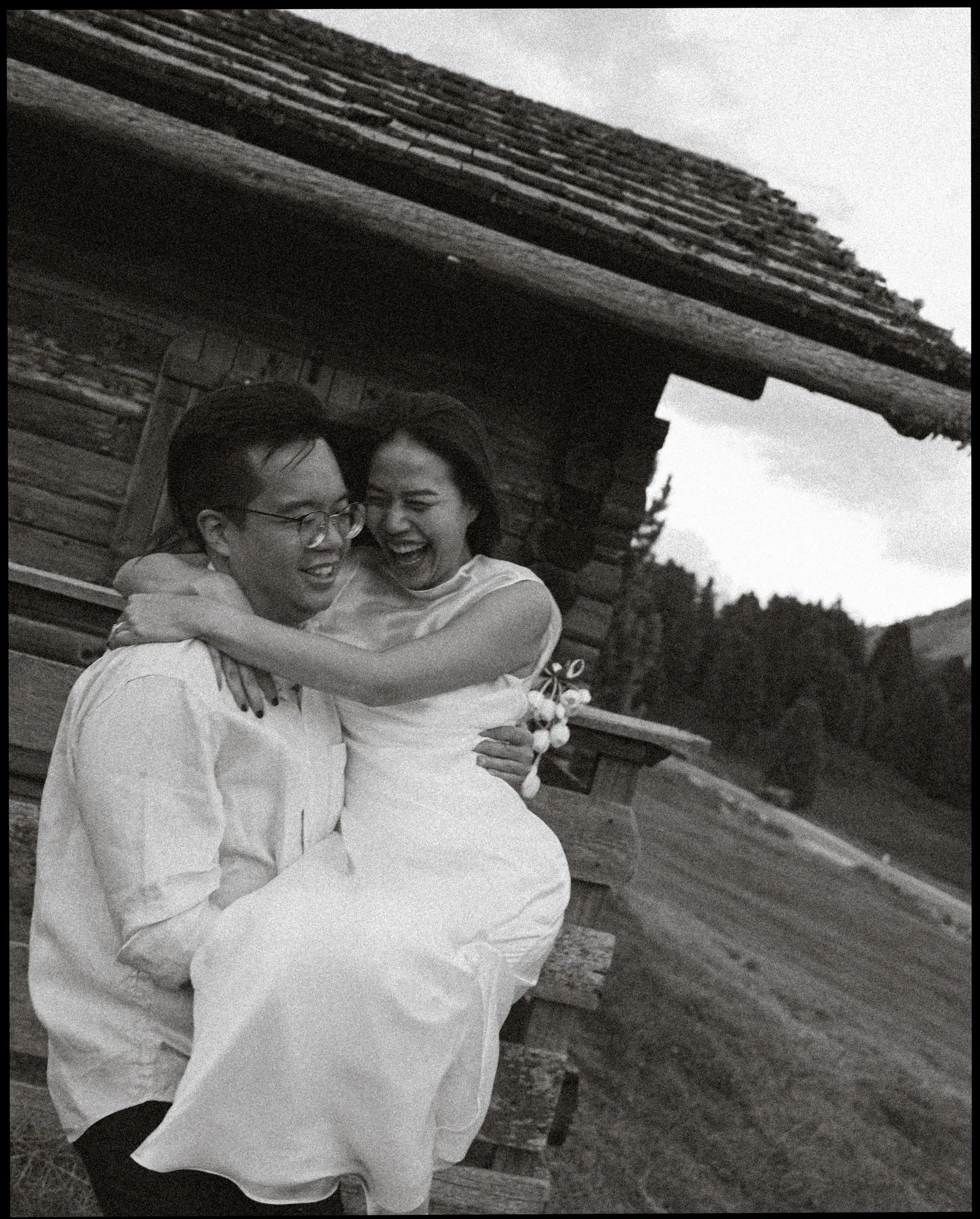 Black and white photo of her sitting on his lap by a hut, both laughing