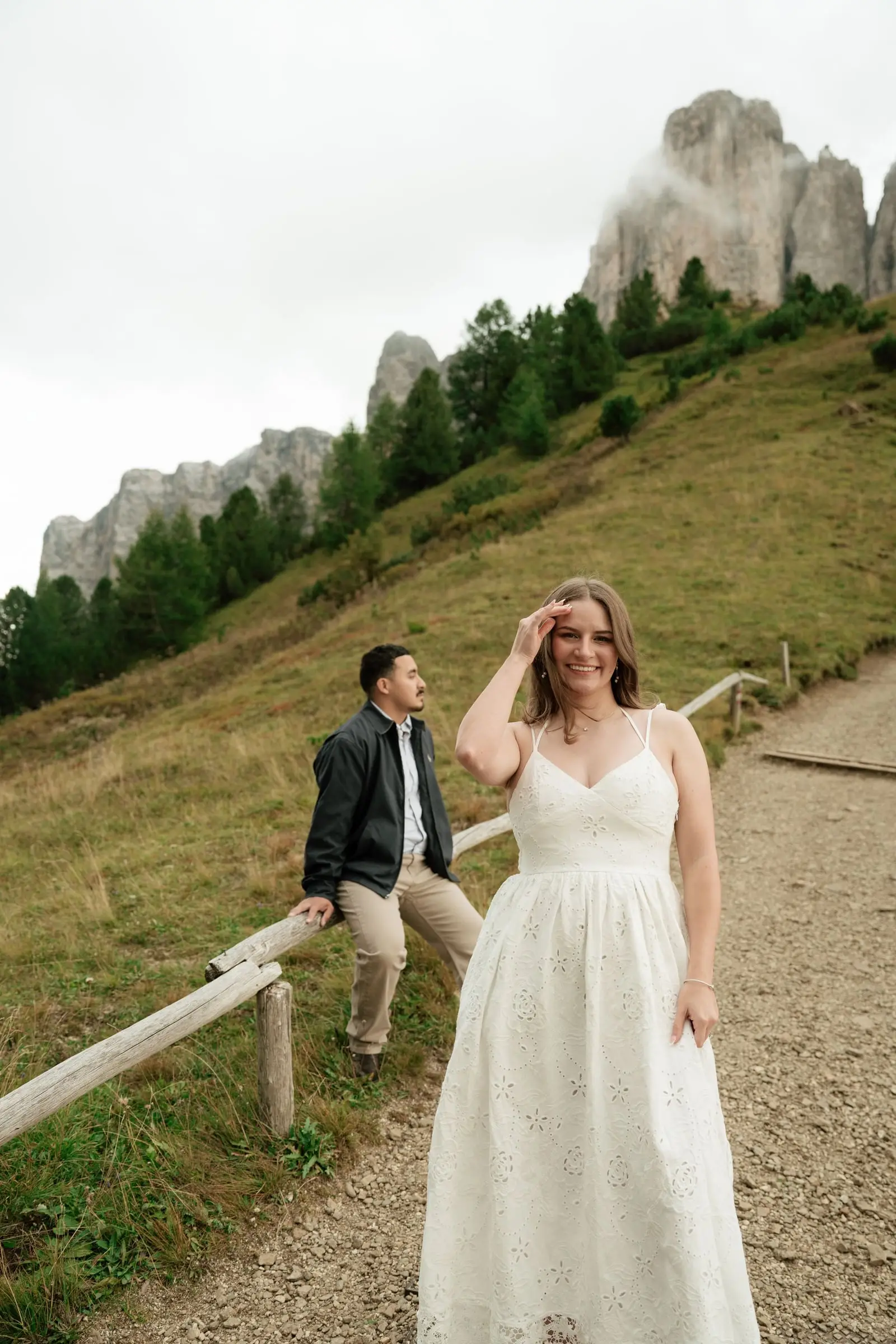 She smiles in a white dress on a mountain path, he sits on a wooden fence