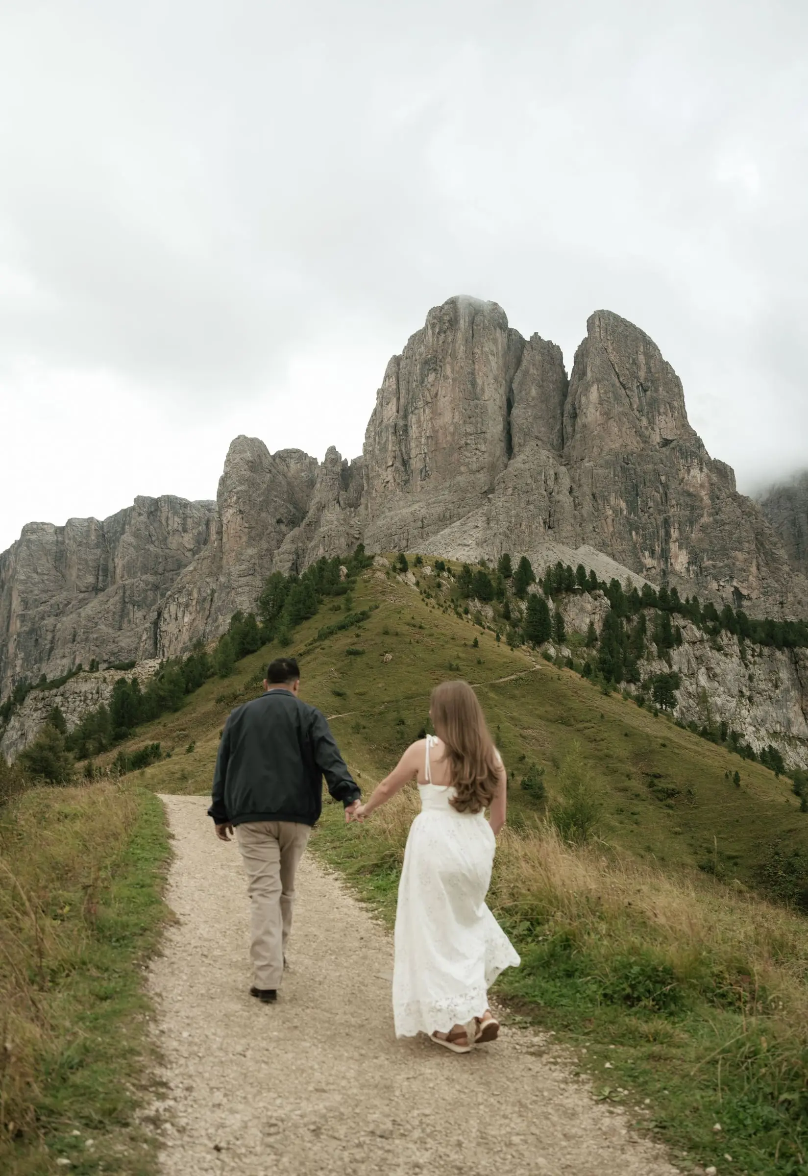 Couple walking hand in hand up a gravel path towards the rock towers of Passo Gardena