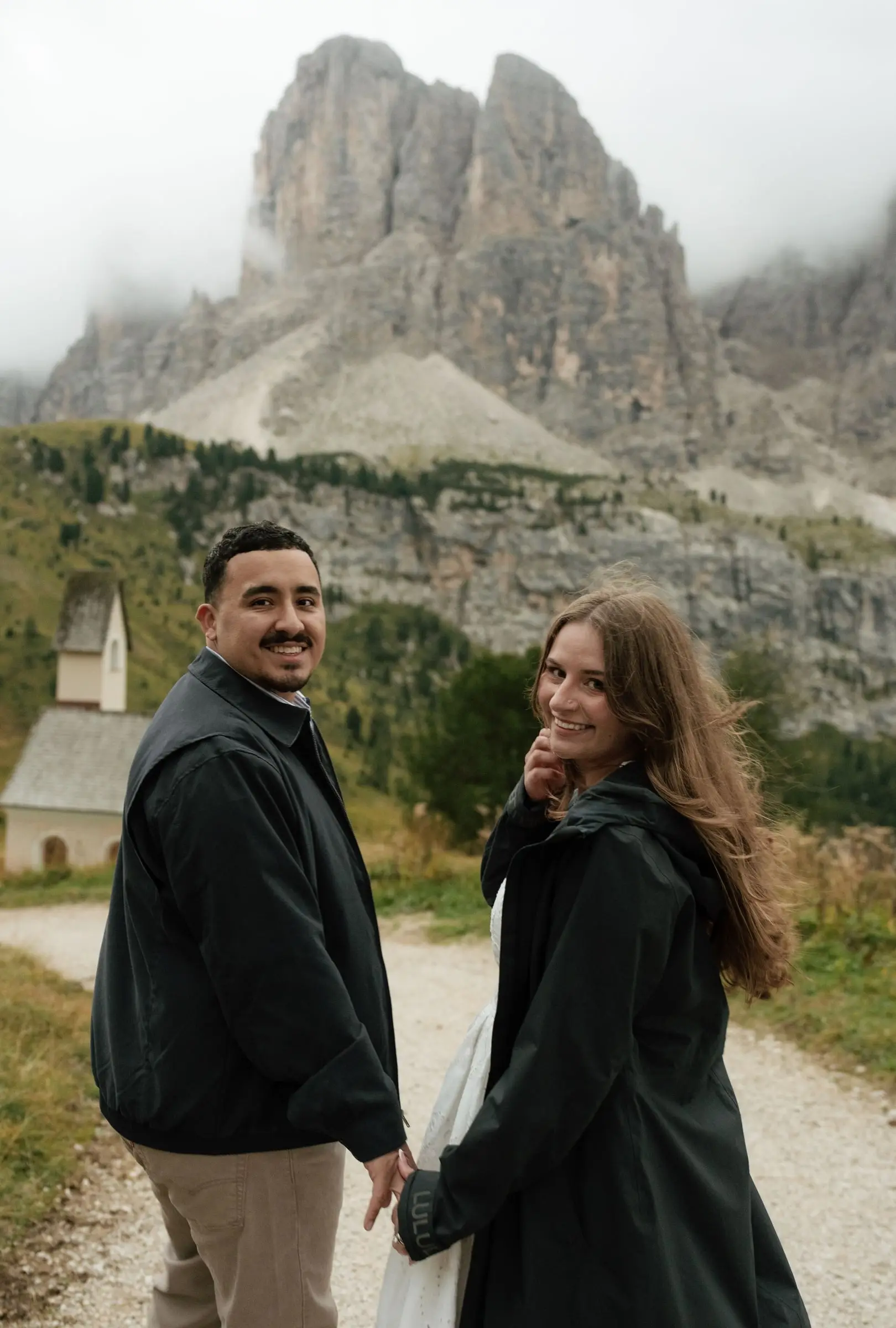 Couple looking back over their shoulders on the path by the little chapel at Passo Gardena