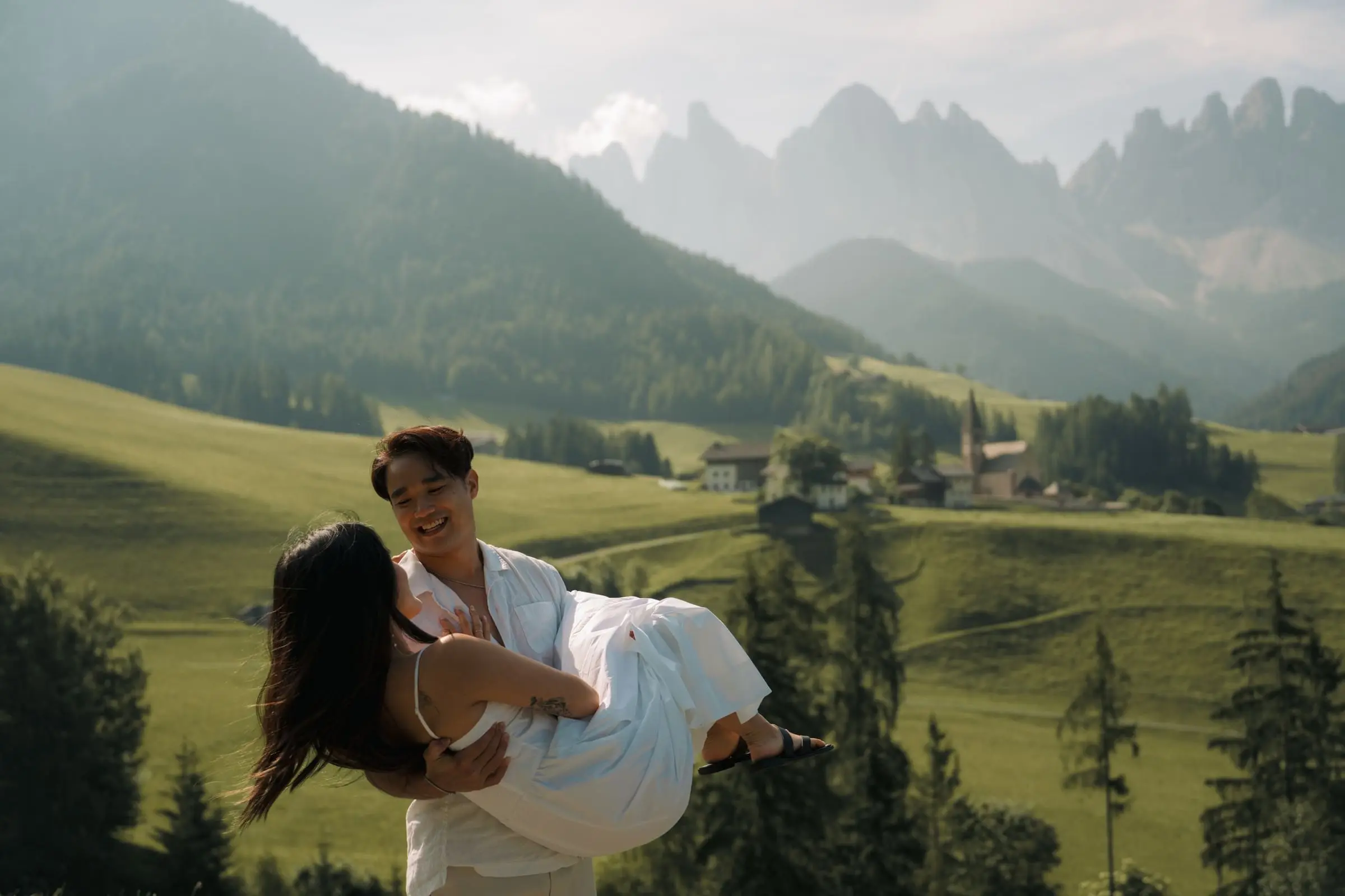 He carries her in his arms above Val di Funes, the Odle peaks in the haze