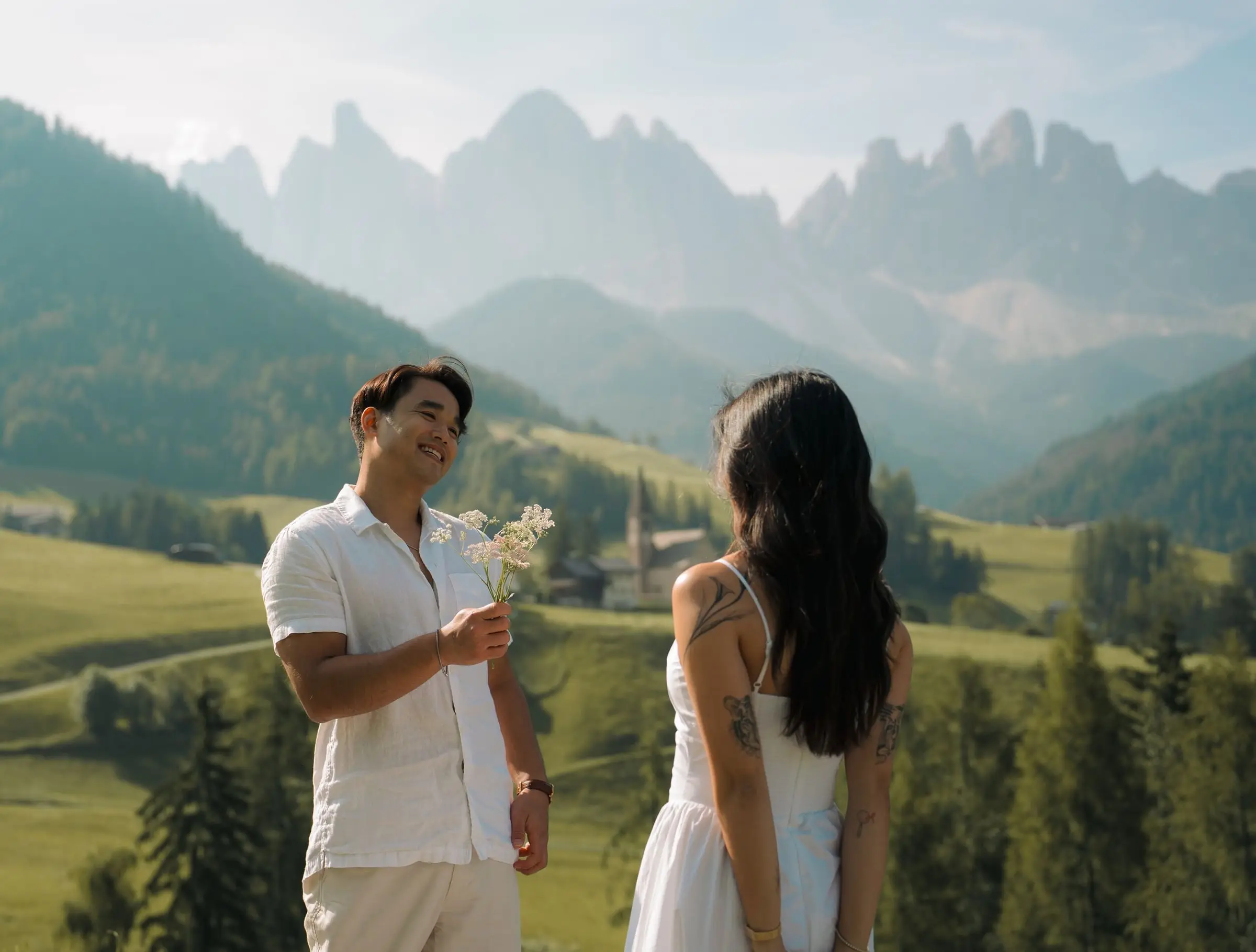 He offers her a small bunch of wildflowers with the Odle peaks behind