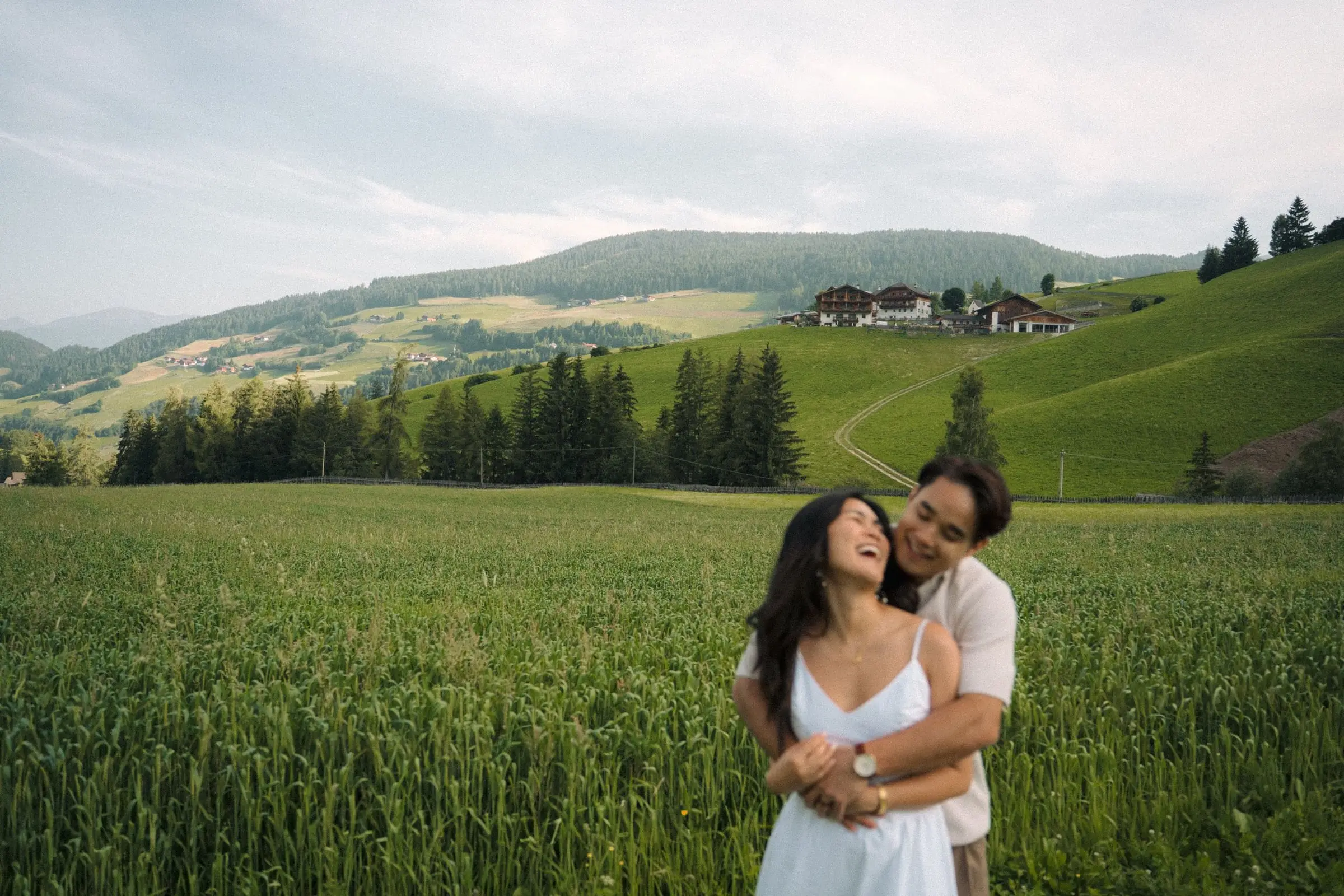 Couple laughing in a green field with farmhouses on the hill