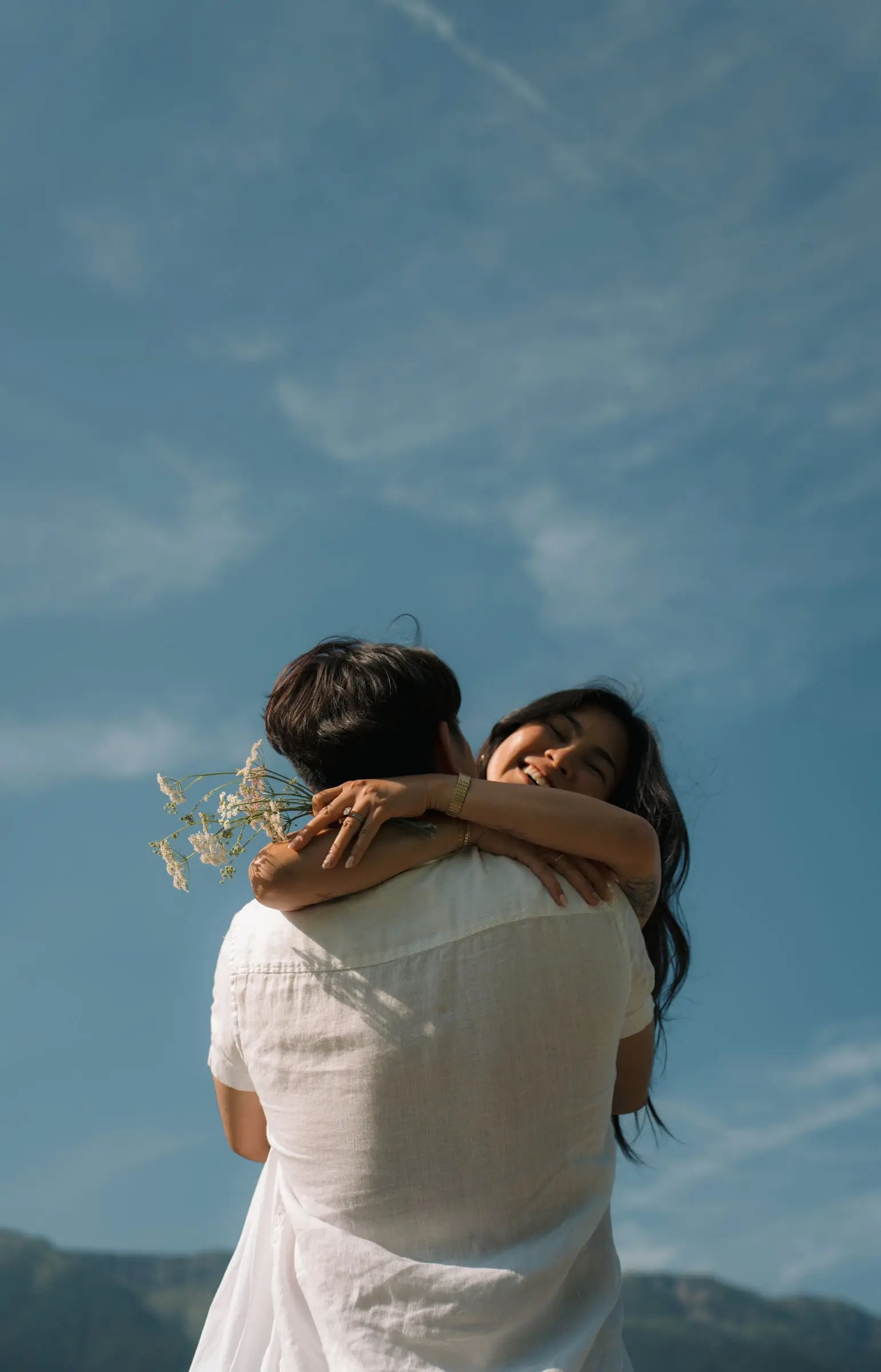 She hugs him with a bunch of wildflowers against the blue sky